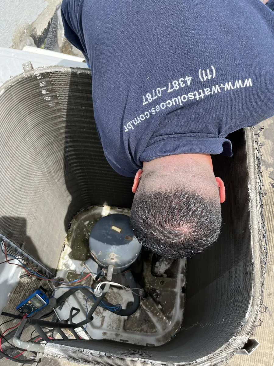 Técnico da Watt Soluções Climatização e Elétrica debruçado na condensadora aberta, com multímetro ao lado, durante conserto de ar condicionado em São Paulo